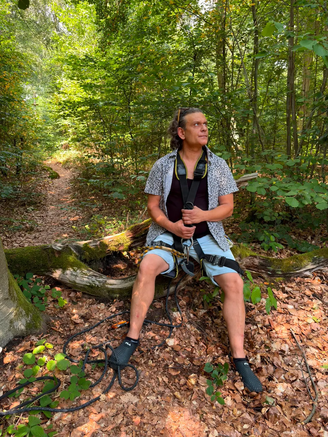 Sven Schwind sitzt mit Klettergurt auf einem umgestürzten Baumstamm und blickt in den Wald